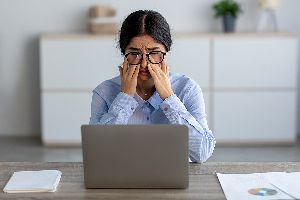 woman sitting a desk in front of her laptop, rubbing her eyes indicating she has a headache.